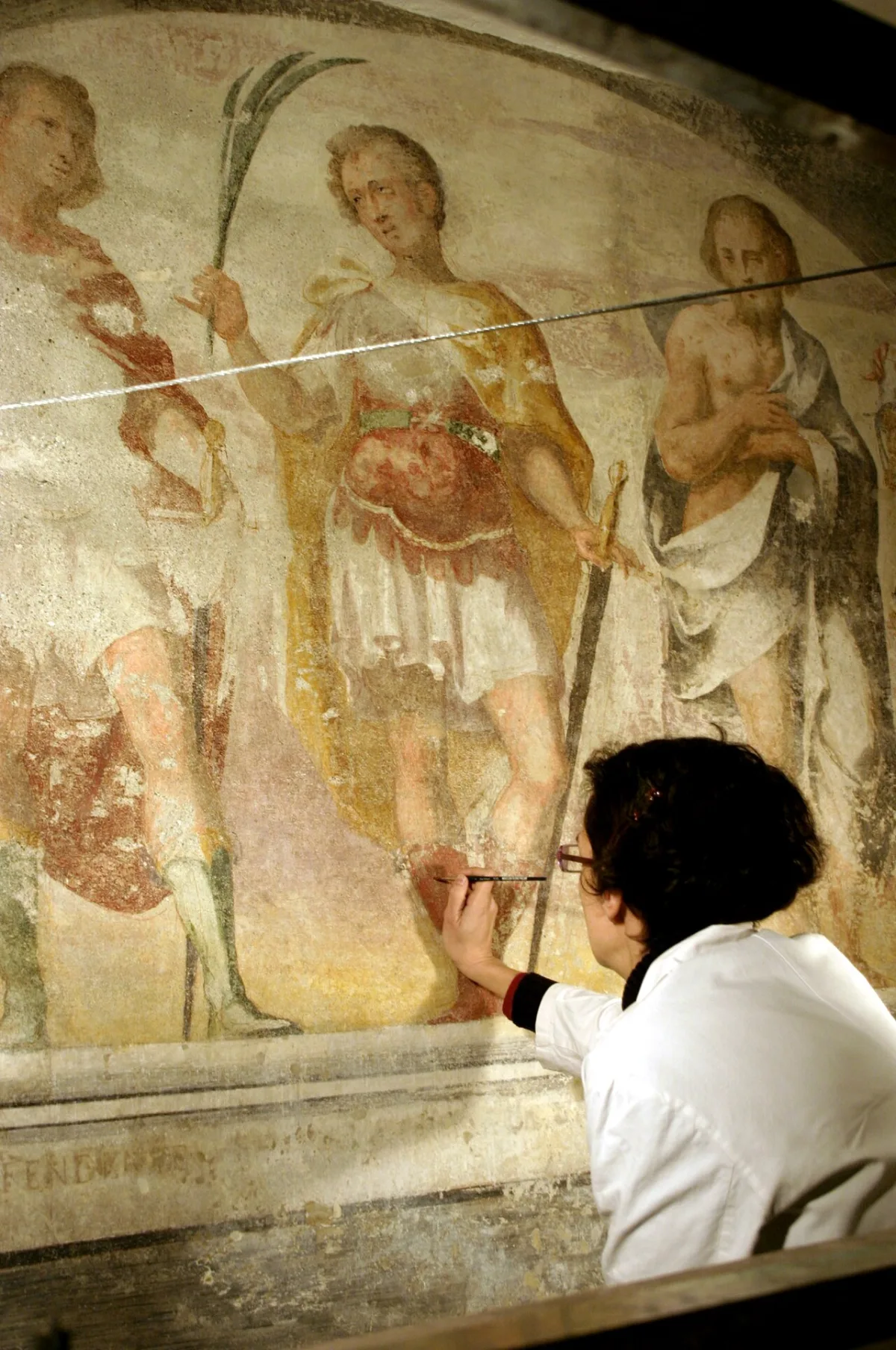 Een restauratrice werkt met een fijn penseel aan een verweerde muurschildering met heiligenfiguren