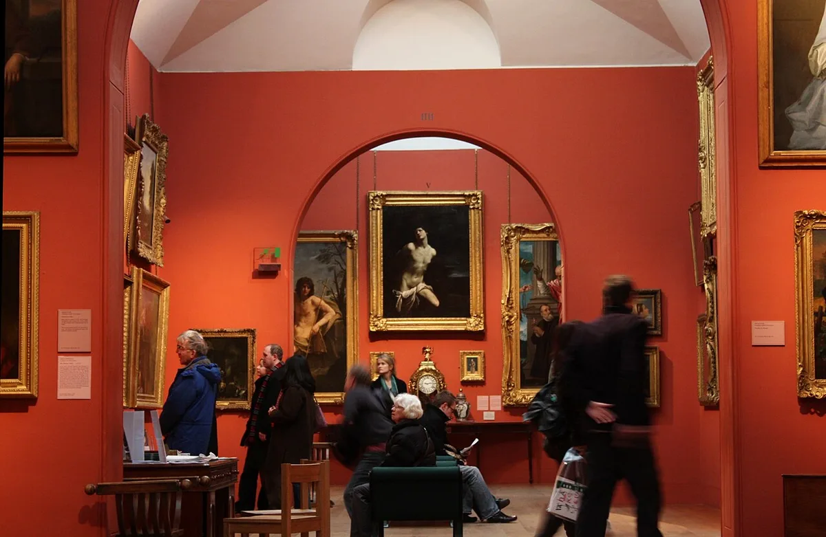Bezoekers in een zaal van de Dulwich Picture Gallery met roodgeschilderde wanden, waar schilderijen in vergulde lijsten in meerdere rijen boven elkaar hangen